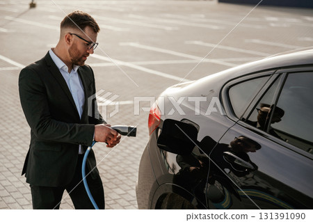 Blue wire for charging is in hand. Businessman in suit is near his black car outdoors Blue wire for charging is in hand. Businessman in suit is near his black car outdoors 131391090