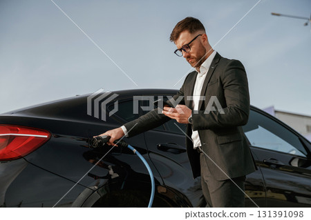 Holding phone in hand while vehicle is charging. Businessman in suit is near his black car outdoors Holding phone in hand while vehicle is charging. Businessman in suit is near his black car outdoors 131391098