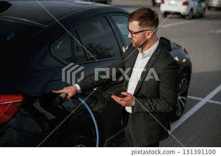 Green energy concept, charging. Businessman in suit is near his black car outdoors Green energy concept, charging. Businessman in suit is near his black car outdoors 131391100