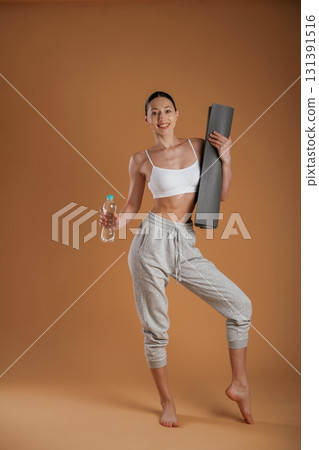 Looking forward, holding bottle of water and mat. Beautiful fitness woman is in the studio Looking forward, holding bottle of water and mat. Beautiful fitness woman is in the studio 131391516