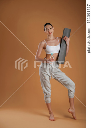 Looking forward, holding bottle of water and mat. Beautiful fitness woman is in the studio Looking forward, holding bottle of water and mat. Beautiful fitness woman is in the studio 131391517