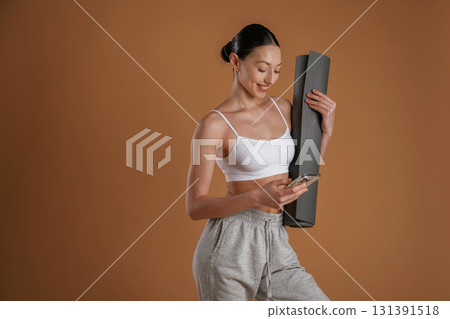 Smiling and holding smartphone with yoga mat. Beautiful fitness woman is in the studio Smiling and holding smartphone with yoga mat. Beautiful fitness woman is in the studio 131391518