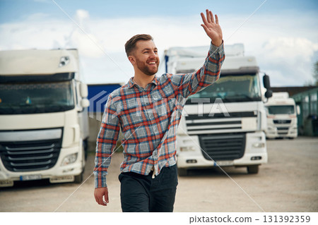 Greetings, hello hand gesture, smiling. Man is standing against trucks outdoors 131392359