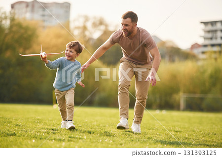 In casual clothes. Running with toy plane. Happy father with son are having fun on the field at summertime In casual clothes. Running with toy plane. Happy father with son are having fun on the field at summertime 131393125