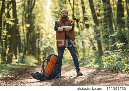Yellow axe in hand, backpack on the ground. Bearded man is in the forest at daytime 131393760