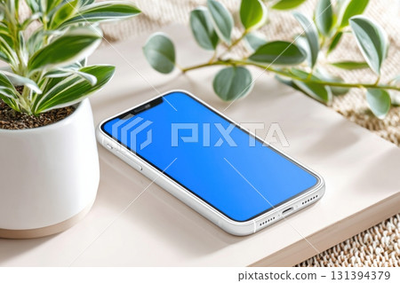 White smartphone with blue app screen rests on a wooden surface beside a potted plant, creating a serene and modern workspace atmosphere with copy space 131394379