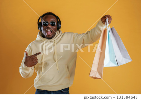 Time for a shopping, with bags, in headphones and sunglasses. Black guy is against yellow background Time for a shopping, with bags, in headphones and sunglasses. Black guy is against yellow background 131394643