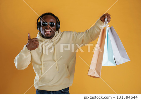 Time for a shopping, with bags, in headphones and sunglasses. Black guy is against yellow background Time for a shopping, with bags, in headphones and sunglasses. Black guy is against yellow background 131394644
