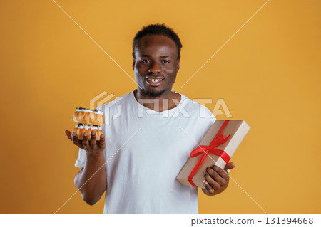 Joyful, holding little cakes and gift box. Black guy is against yellow background Joyful, holding little cakes and gift box. Black guy is against yellow background 131394668