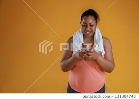 Holding smartphone, towel on the neck. African American woman is in the studio against yellow background Holding smartphone, towel on the neck. African American woman is in the studio against yellow background 131394748