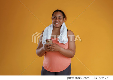 Holding smartphone, towel on the neck. African American woman is in the studio against yellow background Holding smartphone, towel on the neck. African American woman is in the studio against yellow background 131394750