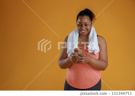 Holding smartphone, towel on the neck. African American woman is in the studio against yellow background Holding smartphone, towel on the neck. African American woman is in the studio against yellow background 131394751