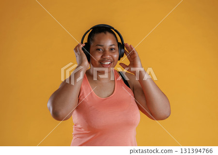 Front view, in headphones, listening to music. African American woman is in the studio against yellow background Front view, in headphones, listening to music. African American woman is in the studio against yellow background 131394766