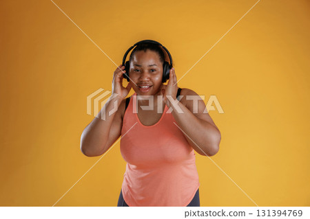 Touching headphones, listening to music. African American woman is in the studio against yellow background Touching headphones, listening to music. African American woman is in the studio against yellow background 131394769