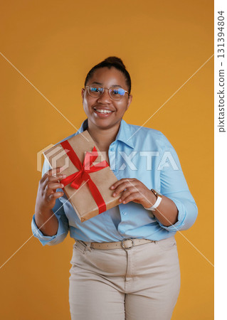 Holidays time, holding gift box. African American woman is in the studio against yellow background Holidays time, holding gift box. African American woman is in the studio against yellow background 131394804