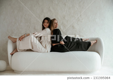 Back to back, sitting on the sofa. Two happy young female best friends are indoors Back to back, sitting on the sofa. Two happy young female best friends are indoors 131395064