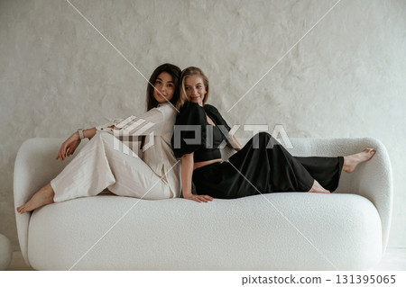 Back to back, sitting on the sofa. Two happy young female best friends are indoors Back to back, sitting on the sofa. Two happy young female best friends are indoors 131395065