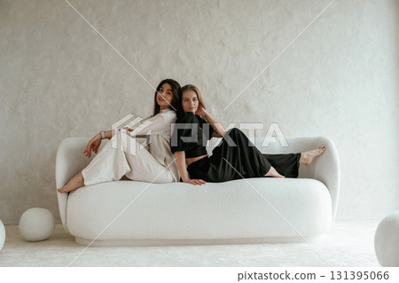 Back to back, sitting on the sofa. Two happy young female best friends are indoors Back to back, sitting on the sofa. Two happy young female best friends are indoors 131395066