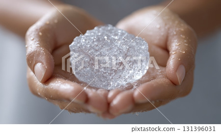 Two graceful hands cup a shiny ice cube, glistening with droplets, creating a fresh and cool appearance 131396031