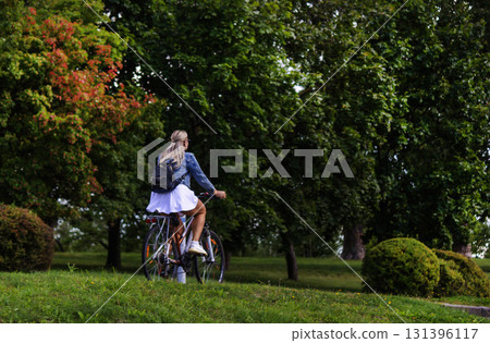 A blonde girl in a white skirt and denim jacket rides a bike 131396117