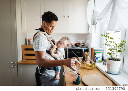 Dad with baby son in carrier in kitchen making tea. Dad with baby son in carrier in kitchen making tea. 131396790