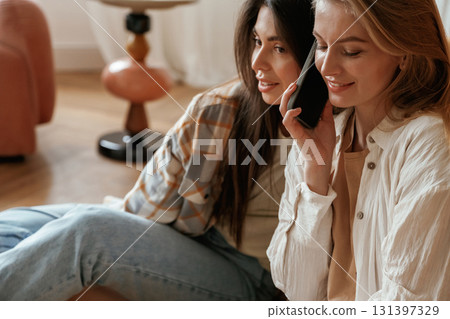 Listening about what woman is talking on phone. Two happy young female best friends are indoors Listening about what woman is talking on phone. Two happy young female best friends are indoors 131397329