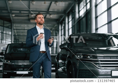 Break, with coffee and smartphone. A businessman is in a car dealership Break, with coffee and smartphone. A businessman is in a car dealership 131397465