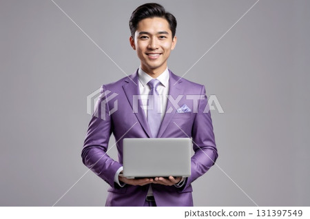 A young man wearing a purple suit stands against a plain background, holding a laptop in his hands. He has a big smile and looks ready to engage, suggesting professionalism and confidence. 131397549