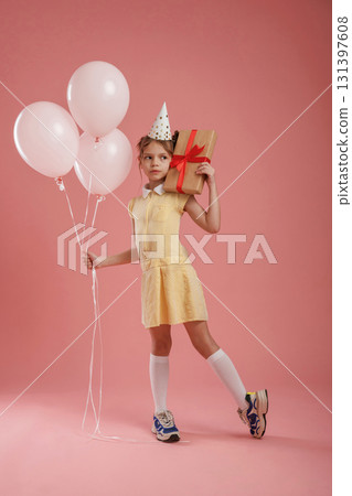 Gift box, birthday hat and balloons. Cute little girl is against pink background Gift box, birthday hat and balloons. Cute little girl is against pink background 131397608