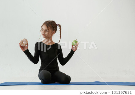 Choice, healthy apple or sweet donut. Little girl in yoga clothes is on the fitness mat Choice, healthy apple or sweet donut. Little girl in yoga clothes is on the fitness mat 131397651