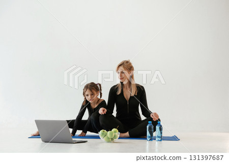 Laptop, apples and bottles of water on the floor. Mother and little daughter are in fitness clothes on yoga mat Laptop, apples and bottles of water on the floor. Mother and little daughter are in fitness clothes on yoga mat 131397687