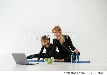 Laptop, apples and bottles of water on the floor. Mother and little daughter are in fitness clothes on yoga mat Laptop, apples and bottles of water on the floor. Mother and little daughter are in fitness clothes on yoga mat 131397688