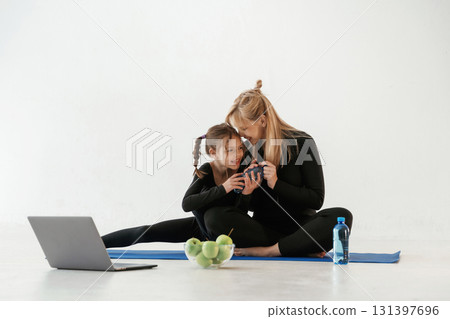 Silver colored laptop, apples and water. Mother and little daughter are in fitness clothes on yoga mat 131397696