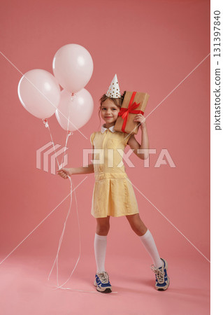 Gift box, birthday hat and balloons. Cute little girl is against pink background Gift box, birthday hat and balloons. Cute little girl is against pink background 131397840