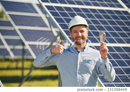 Smiling and holding bulb. Engineer with photovoltaic solar panels outdoors at daytime 131398449