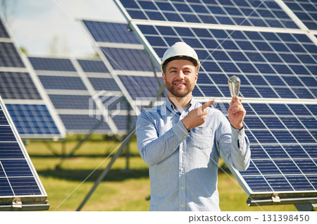 Smiling and holding bulb. Engineer with photovoltaic solar panels outdoors at daytime Smiling and holding bulb. Engineer with photovoltaic solar panels outdoors at daytime 131398450