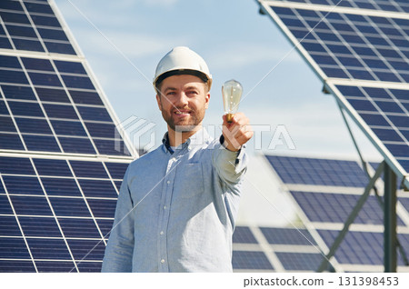 Smiling and holding bulb. Engineer with photovoltaic solar panels outdoors at daytime Smiling and holding bulb. Engineer with photovoltaic solar panels outdoors at daytime 131398453