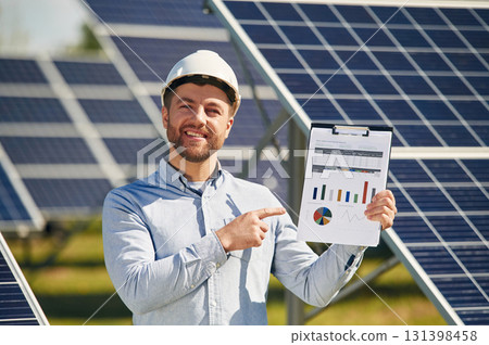 Pointing to the notepad with diagrams and graphs. Engineer with photovoltaic solar panels outdoors at daytime 131398458