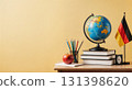 A classroom desk displays a globe representing the world, stacked books, colored pencils in a holder, and a red apple beside a German flag on a yellow background. 131398620