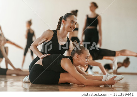 Woman with pigtail is helping little girl. Group of female kids practicing athletic exercises together indoors 131399441