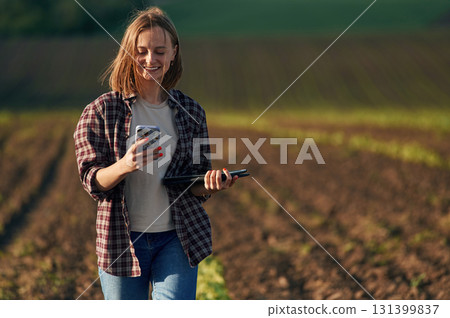 Smartphone and tablet in hands. Young woman is on the beautiful agricultural field at daytime Smartphone and tablet in hands. Young woman is on the beautiful agricultural field at daytime 131399837