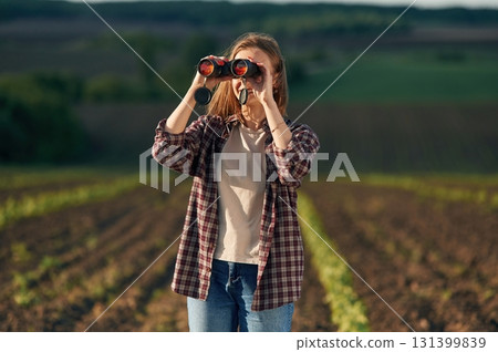 Looking through the binoculars. Young woman is on the beautiful agricultural field at daytime Looking through the binoculars. Young woman is on the beautiful agricultural field at daytime 131399839