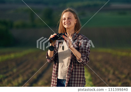 Looking through the binoculars. Young woman is on the beautiful agricultural field at daytime Looking through the binoculars. Young woman is on the beautiful agricultural field at daytime 131399840