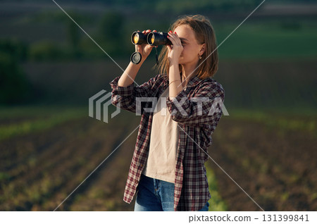 Looking through the binoculars. Young woman is on the beautiful agricultural field at daytime Looking through the binoculars. Young woman is on the beautiful agricultural field at daytime 131399841