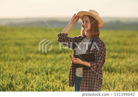 Positive mood, with straw hat and with documents. Young woman is on the beautiful agricultural field at daytime 131399842