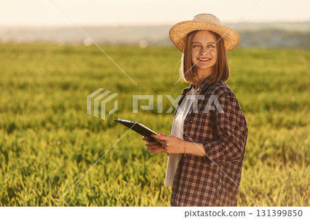 Front view, standing and holding documents. Young woman is on the beautiful agricultural field at daytime Front view, standing and holding documents. Young woman is on the beautiful agricultural field at daytime 131399850
