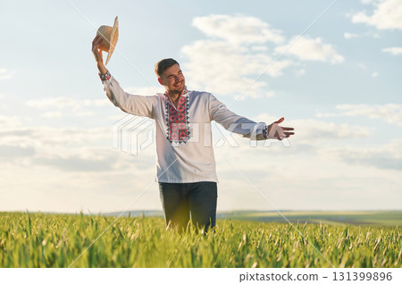 Taking off straw hat. Man in Ukrainian embroidery shirt is on the agricultural field at summer daytime 131399896