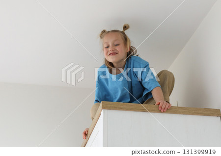 Funny kid girl sitting on a high cabinet. Child 6 years old at home 131399919
