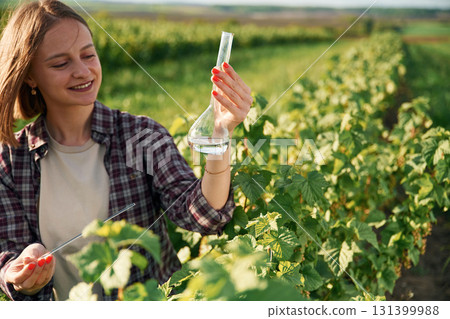 With test tube, doing research. Young woman is on the beautiful agricultural field at daytime With test tube, doing research. Young woman is on the beautiful agricultural field at daytime 131399988