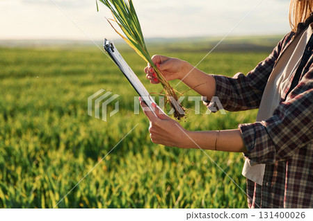 Side view. With notepad and plant. Young woman is on the beautiful agricultural field at daytime Side view. With notepad and plant. Young woman is on the beautiful agricultural field at daytime 131400026